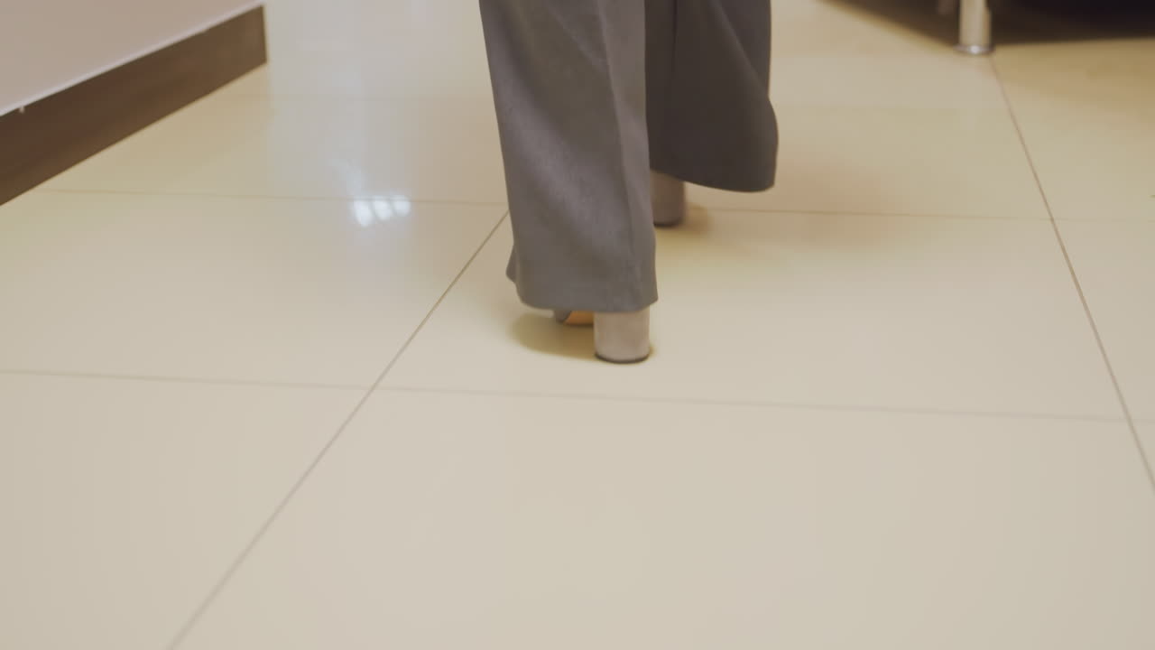 Close-up of businesswoman's feet walking in gray heels on office floor with geometric tile pattern, confident professional stride, modern office corridor, stylish and formal footwear