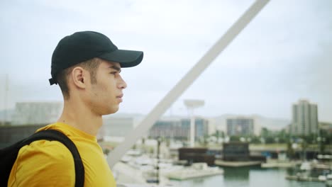 Young-Attractive-Trendy-young-Man-wearing-a-cap-and-a-backpack-walking-on-a-bridge-on-a-sunny-day-with-an-urban-city-background