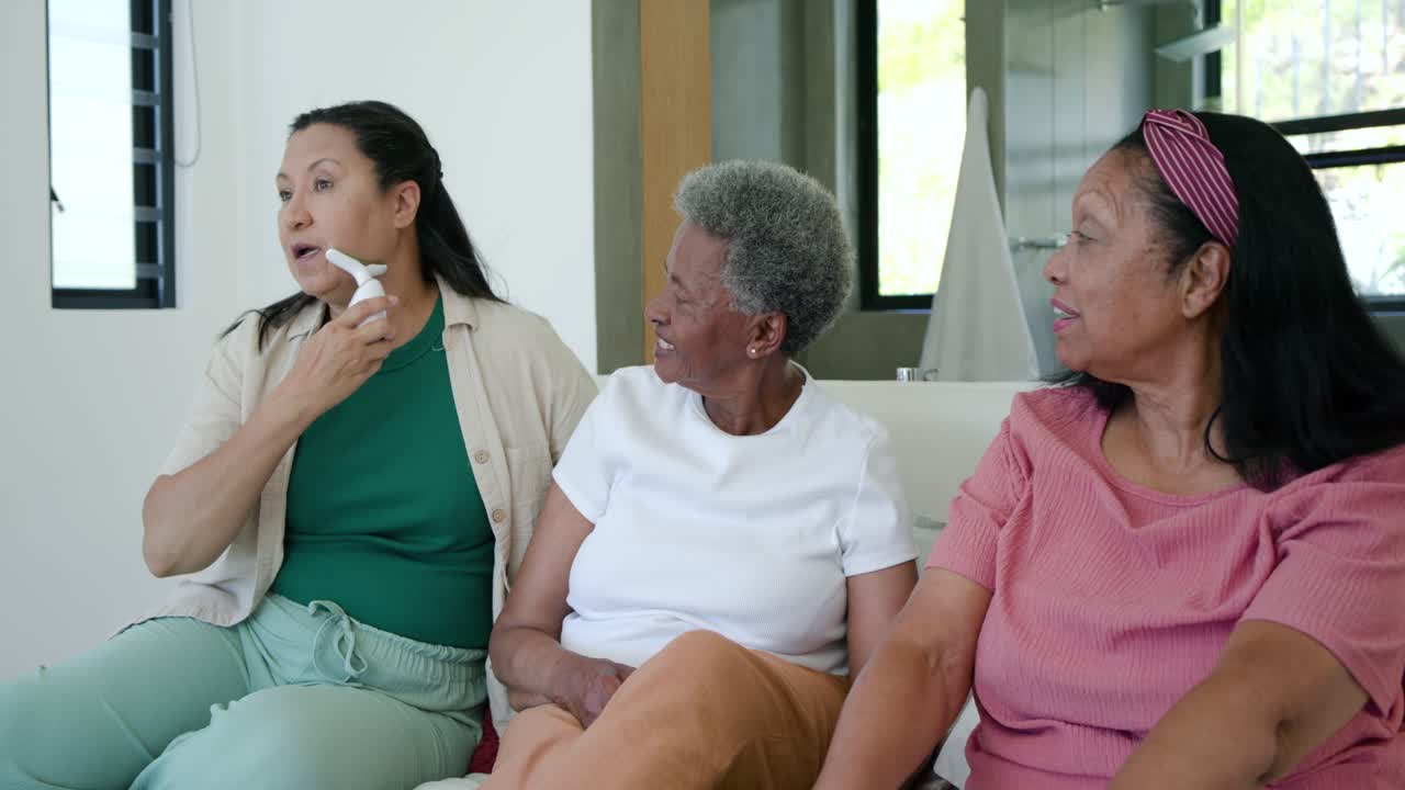African American female relatives watching older relative showing white device on sofa for seniors