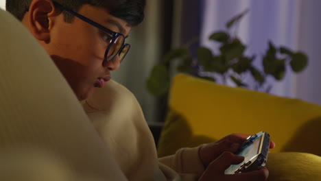 young boy sitting on sofa at home playing game or streaming onto handheld gaming device at night 3