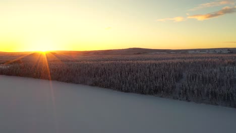 Aerial-view-of-sunset-over-snowy-frozen-lake-in-deep-forest-with-hills-and-mountains