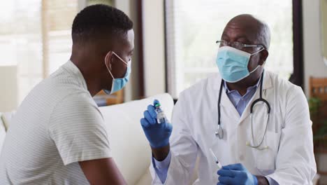 African-american-senior-male-doctor-giving-covid-vaccine-to-male-patient-in-home,-wearing-face-masks