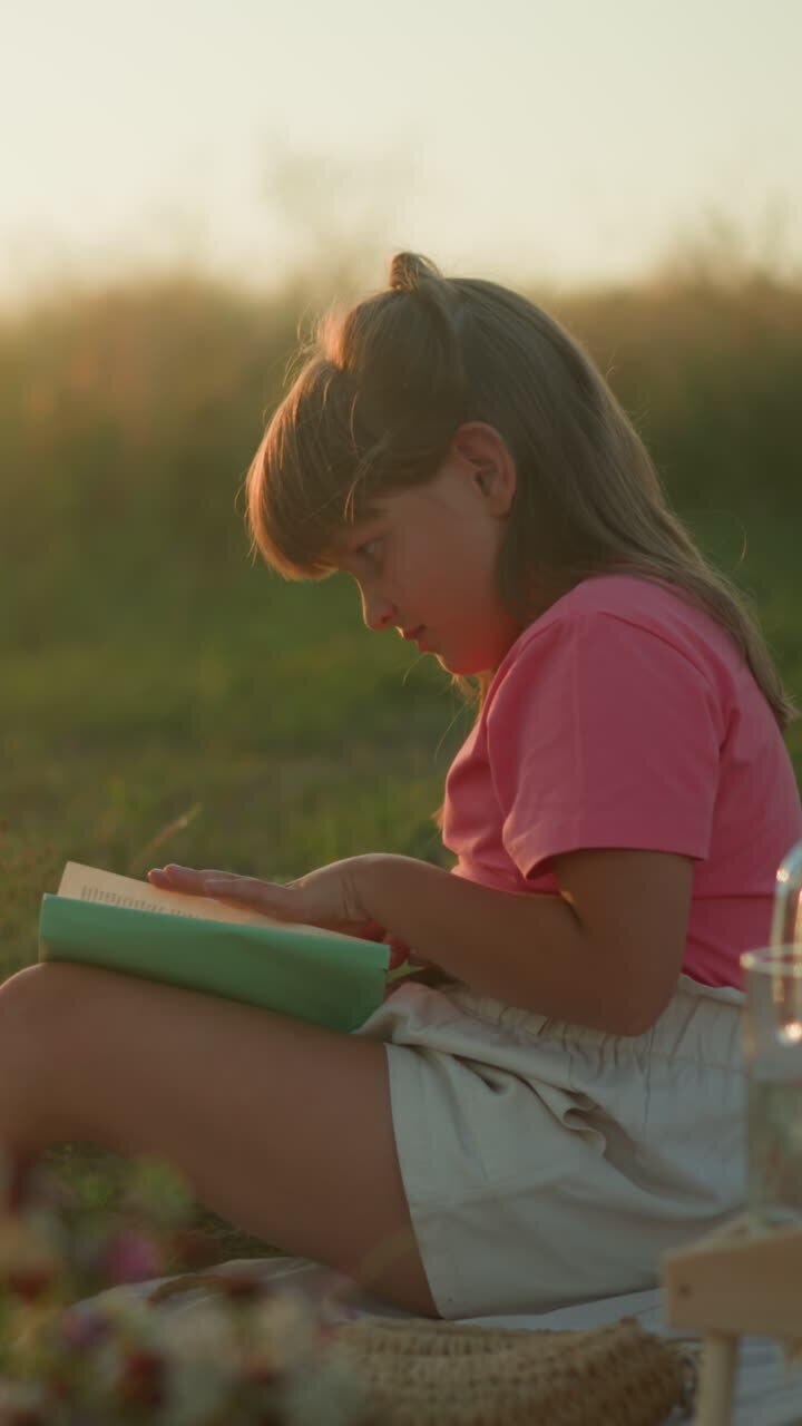 Little girl sitting outdoors during golden hour reading book and turning page, surrounded by picnic setup with fruits, croissant, and bubbles gently floating in air, sunlight illuminating background
