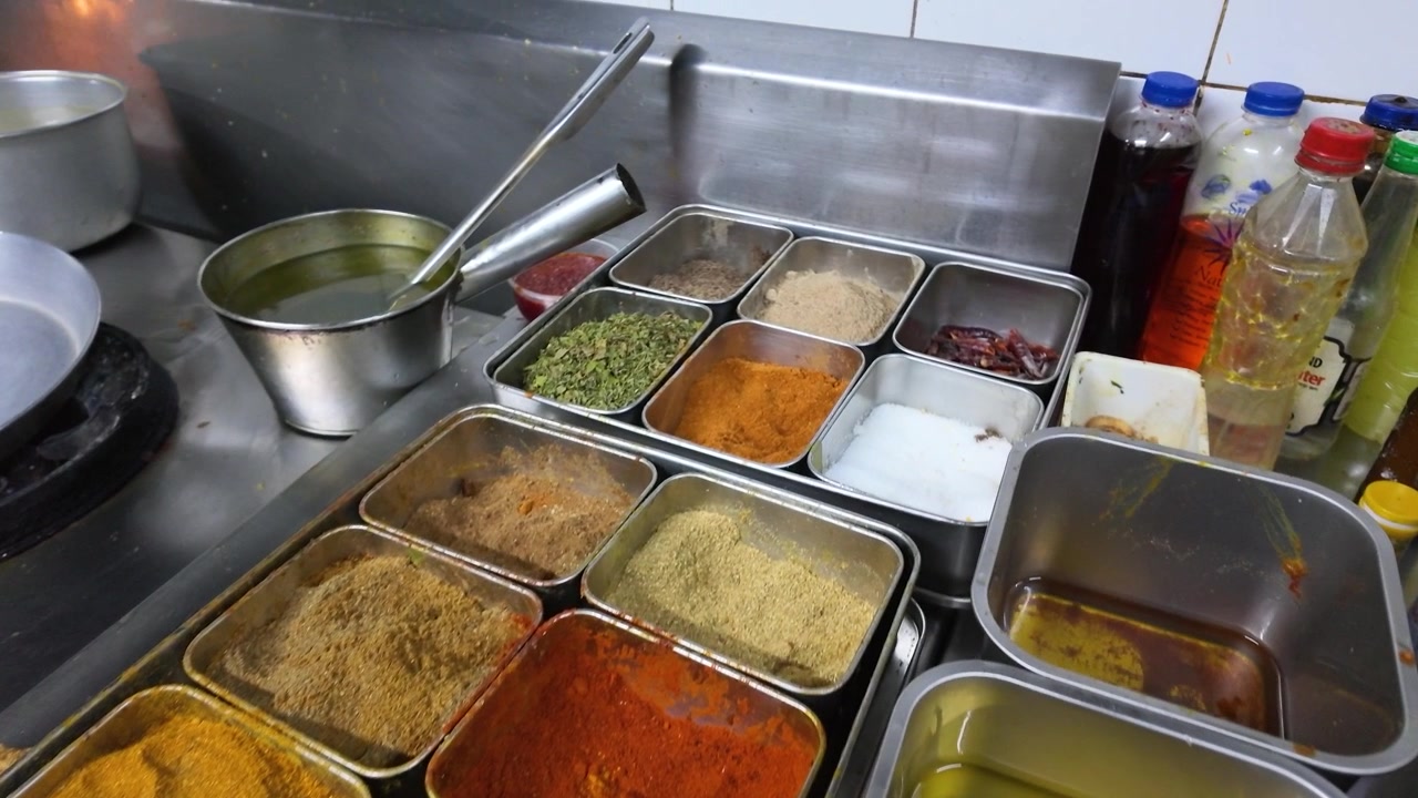 Ripresa panoramica ravvicinata di spezie e polveri colorate in contenitori di acciaio inossidabile all'interno di una cucina professionale.