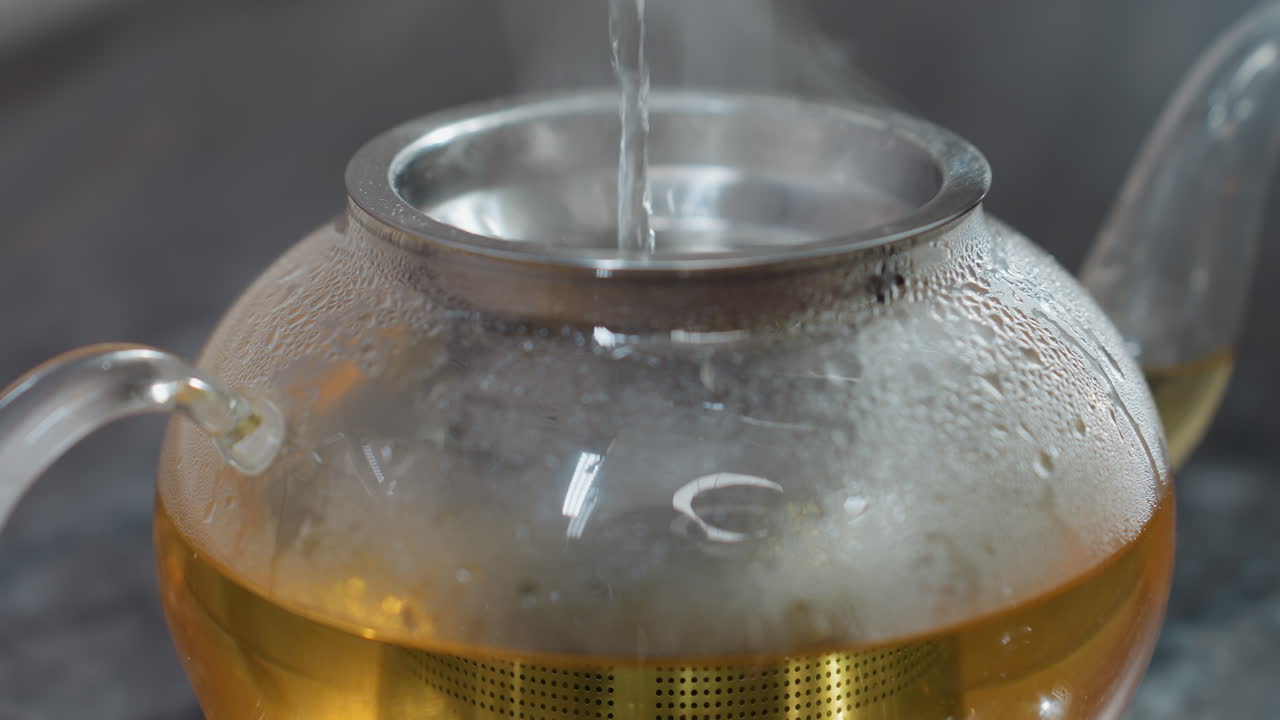 Close up of pouring hot water into glass kettle as steam rises, condensation forming on surface, golden herbal tea brewing inside with stainless steel filter
