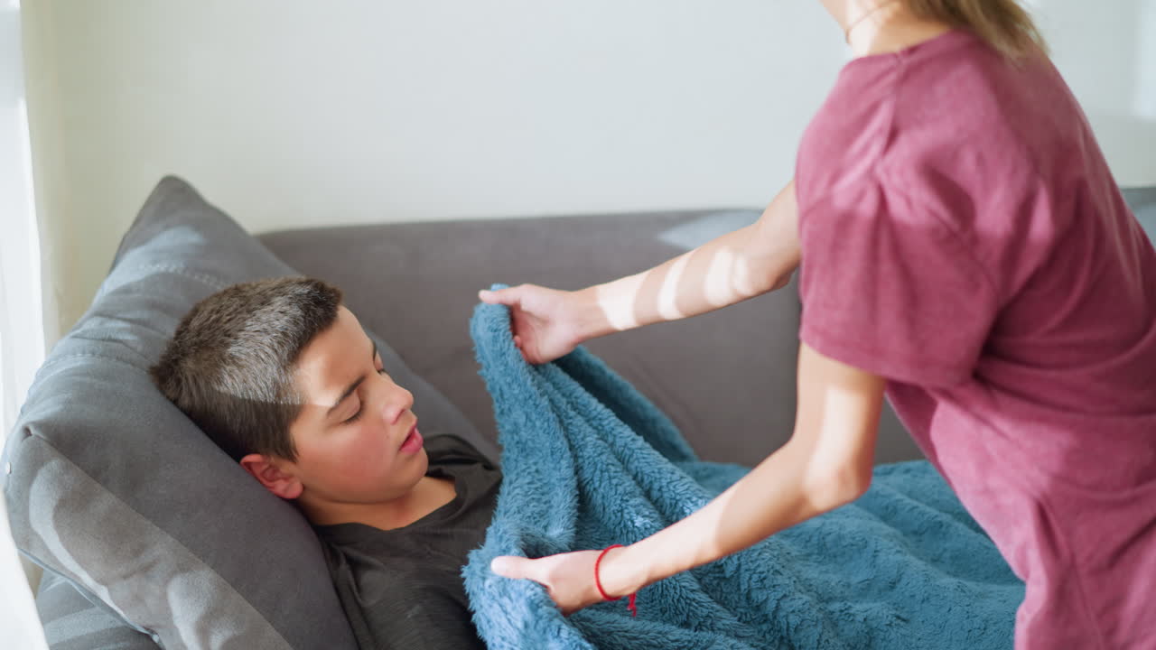Close-up of boy sleeping on couch with blue blanket, sister coming to cover him carefully with soft blanket, shadow of her hand cast on blanket