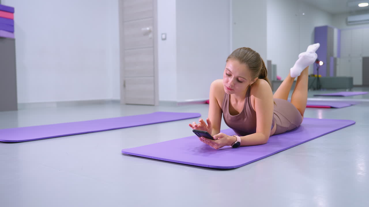 mujer joven yace en la alfombra de yoga, centrado en el teléfono, levantando las piernas ligeramente, el fondo cuenta con grandes espejos y gabinetes de almacenamiento en un estudio de fitness moderno