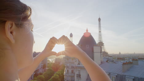 young woman hands making heart shape gesture holding sun flare enjoying romantic travel vacation in paris france looking at beautiful eiffel tower