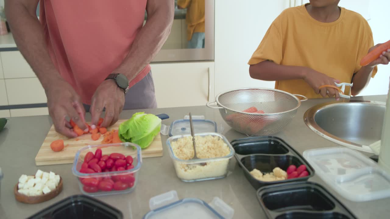 Afroamerikanischer Vater und Sohn schneiden Karotten und schälen an der Spüle, portionieren Mahlzeiten zur Lagerung.