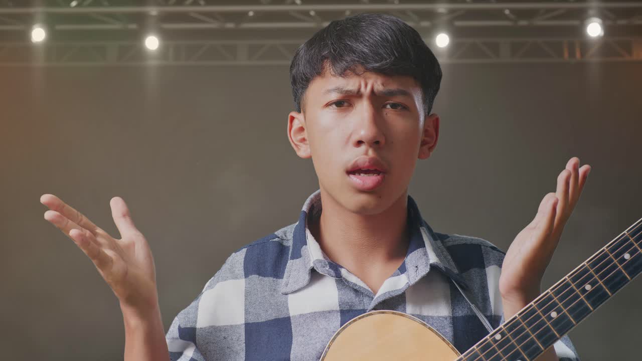 기타를 들고 있는 아시아 청소년의 클로즈업은 당황스러운 몸짓을 보여주고 무대에서 무엇을 말하고 있습니다.