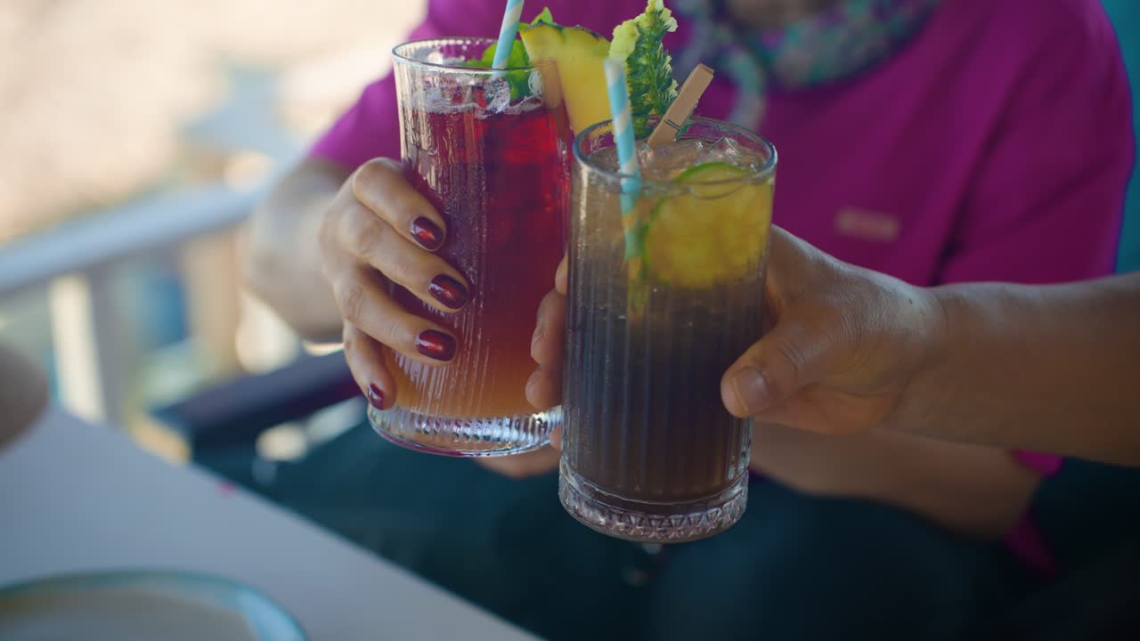 Close-up slow motion of senior couple clinking garnished tropical cocktails with pineapple, colorful straws and ice while relaxing on summer vacation