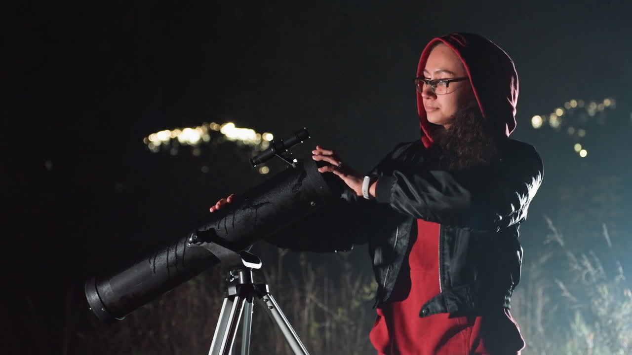 Un osservatore con un telescopio che scruta il cielo notturno, regola il telescopio, indossa una felpa rossa con cappuccio e una giacca di pelle, si trova accanto a un riflettore montato su un treppiede, regola l'oculare e allinea le ottiche, sullo sfondo le luci fioche della città.
