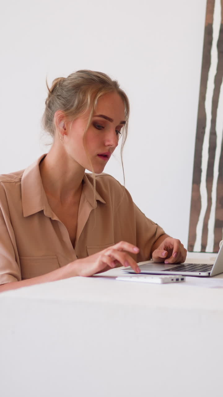 Young blonde woman with hair bun in beige shirt sits at white table making calculations on modern laptop and numeric keyword in light room slow motion