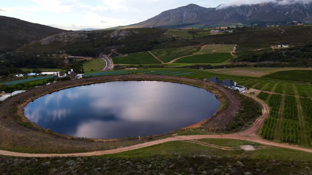 Perfectly round, man made dam on a wine farm reflects the cloudy sky like a mirror, in-between the vineyards with mountains in the distance