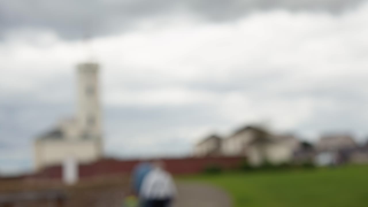 Person walks toward distant lighthouse on grassy path, overcast sky, handheld camera, soft focus