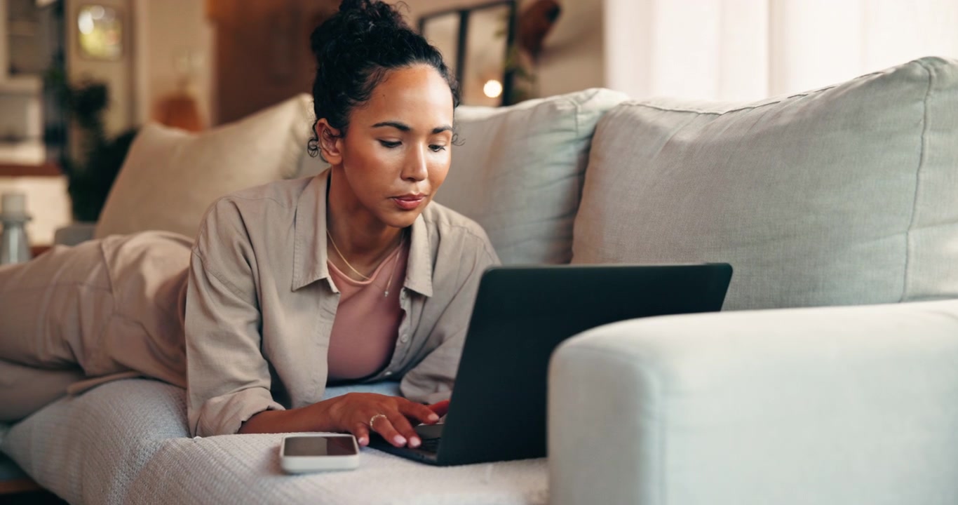 Ung kvinde arbejder på bærbar computer, mens hun ligger i sofaen derhjemme