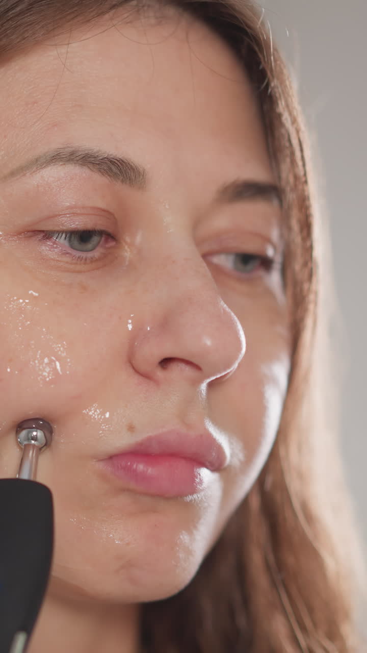 얼굴 리프팅을 위해 미세 전류 절차를 수행하는 여성. 예쁜 갈색 여성은 집 화장실에서 얼굴 위에 전기 장치로 손을 고 느린 동작을합니다.