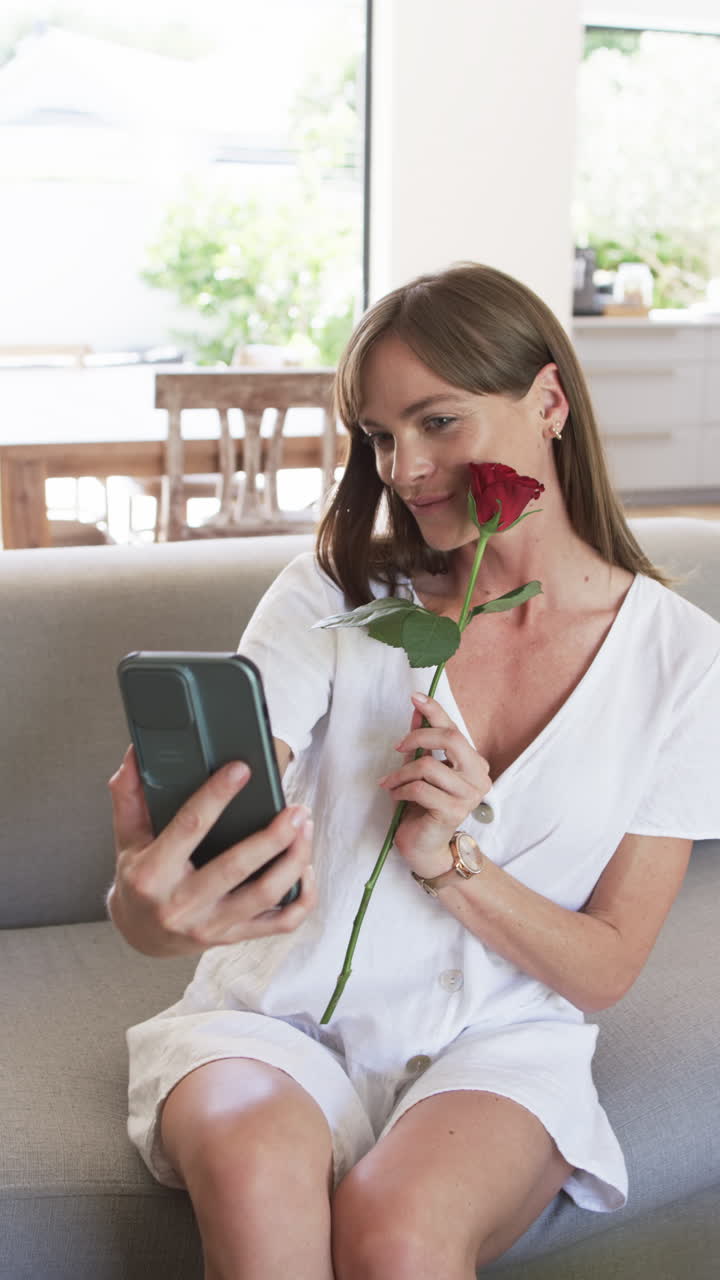 Vertical video: Caucasian woman holding red rose, smiling at phone at home