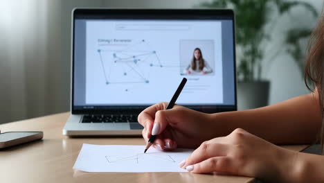 person drawing on paper while attending an online meeting on a laptop