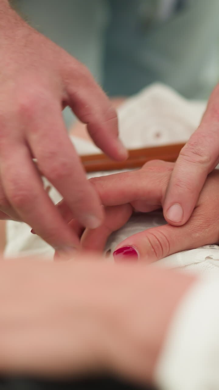 A close-up shot of a man tenderly positioning a woman's hands on her lap, highlighting her vibrant red nails and a distinctive ring, all set against the calming backdrop of a tranquil green field