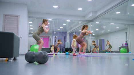 gym session showing two women performing squats with resistance bands, watched by a toddler with a band on her leg, the setting includes a mirror reflecting their movements and gym balls around