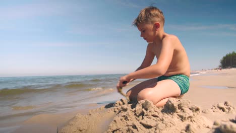 young boy in swimwear plays at the beach and digs a path into the sand with shovel