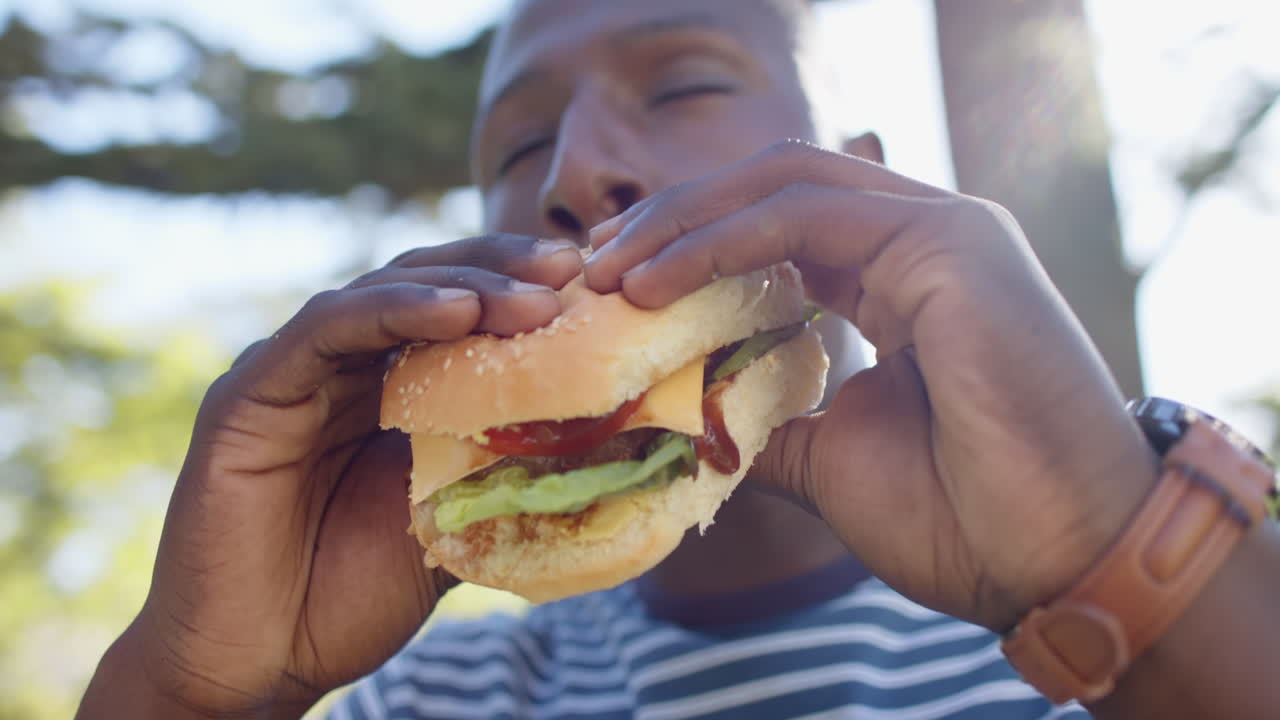 Eating burger, african american man enjoying outdoor hangout with friend in park