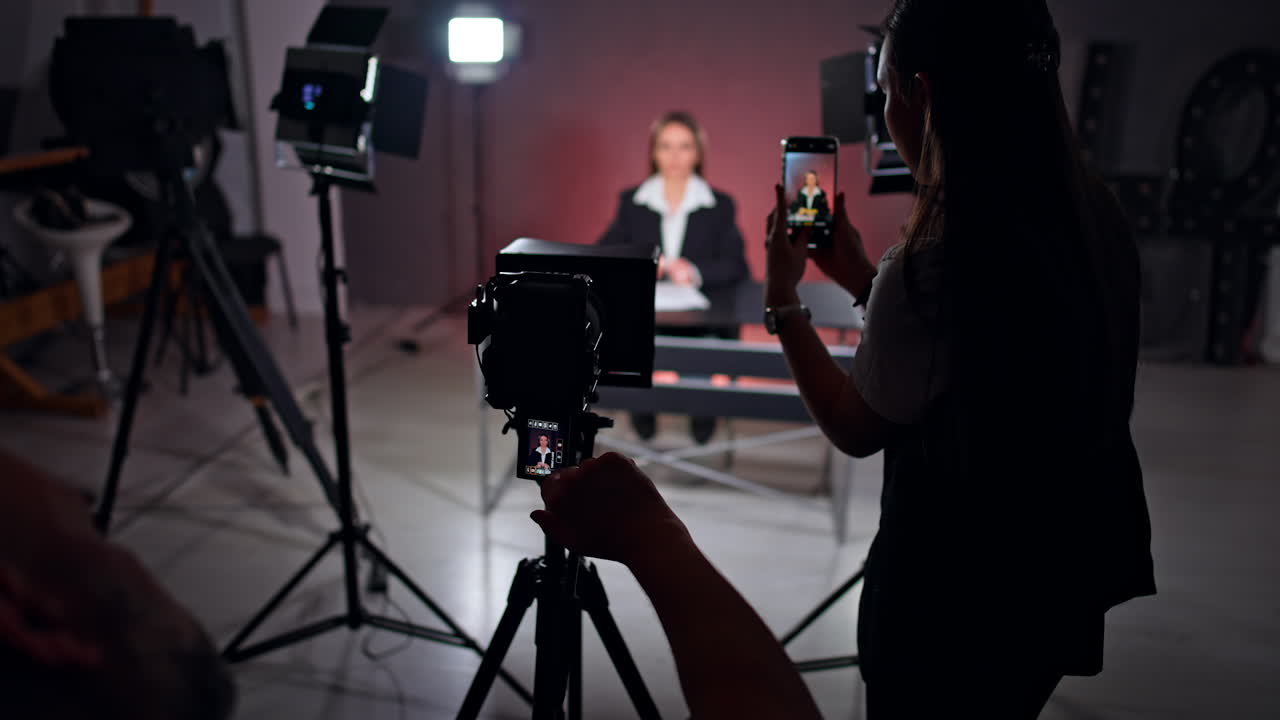 Woman in black jacket is filmed on the professional camera and smartphone. Blog content creation in studio. TV program recording.