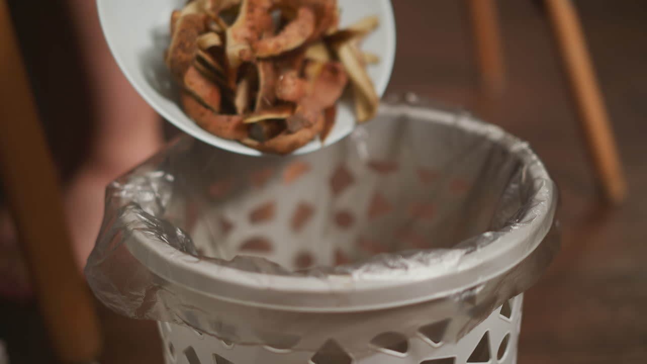 Close up of ash colored patterned waste bin lined with plastic bag as person seated on wooden chair disposes potato peels into it, with visible bare feet on wooden floor