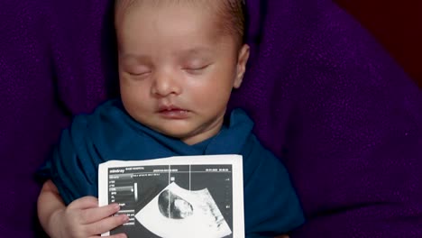 newborn baby holding ultrasound copy in hand and sleeping at mother palm in baby wrap