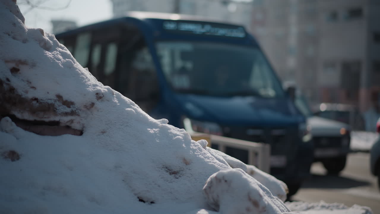 Snow heap beside city road in winter, cars drive past along lane, blurred urban buildings in background, bright sun glints on windshields, gritty plowed mound frames traffic movement