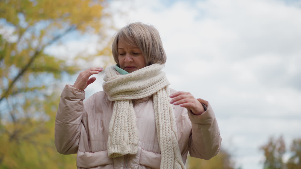 close up of woman adjusting chunky knit neck scarf around neck while standing in autumn garden with golden foliage blurred background wearing pink coat