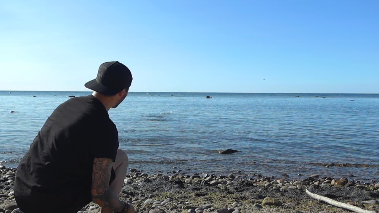 Slow motion footage of a caucasian male throwing and skipping a rock or a pebble on blue ocean sea water surface, creating waves and water ripples during summer sunny day at a rocky beach shoreline