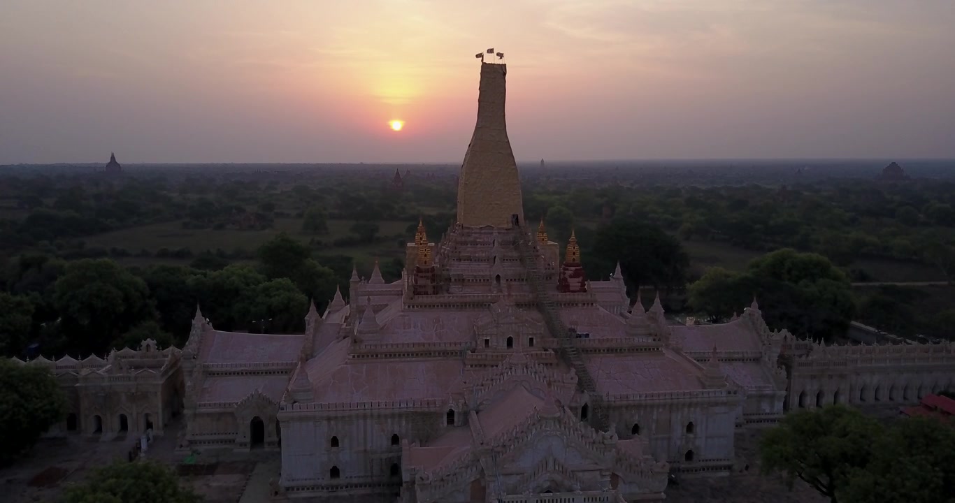 Un drone avanza lentamente verso il Tempio Ananda imbiancato mentre il sole tramonta dietro la sua guglia dorata, nella piana degli antichi templi di Bagan. Vista aerea.