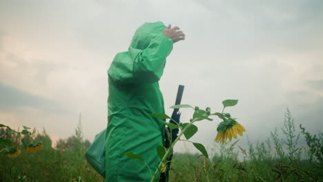 mujer camina más allá del girasol, se quita la capucha del impermeable mientras sostiene un paraguas, observando de cerca la flor mientras caen gotas de agua, en un campo verde brumoso que se extiende hacia el horizonte, enmarcado por arbustos