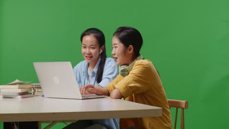 asian woman student teaching her friend on a laptop while sitting on a table in the green screen background classroom