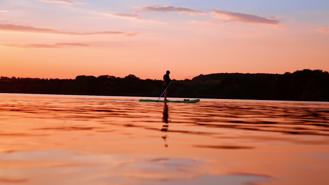 Sup boarding on the river at sunset time. Lonely silhouette of a sportsman rowing on the board by the waterscape colored orange.