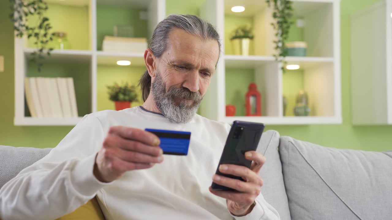 Anziano felice che fa acquisti online con il suo smartphone a casa, pagando con carta di credito