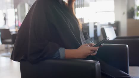 happy caucasian female client in cape at hair salon using smartphone, in slow motion