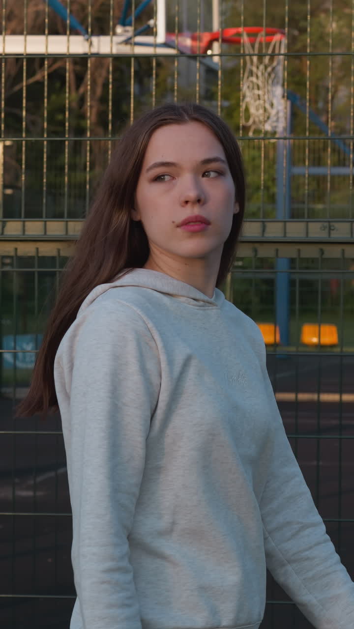 dama con capucha pasa por la valla de un campo de deportes. caminando y relajándose después de la clase de fitness en el aire fresco al atardecer. sensación de vacío y soledad