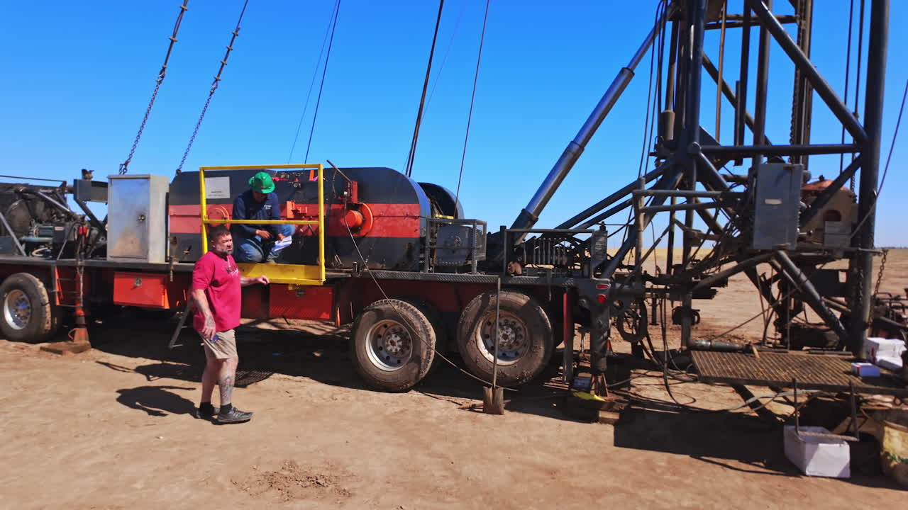 Approaching the vehicle with the derrick for oil production. Few men work at equipment for drilling resources.