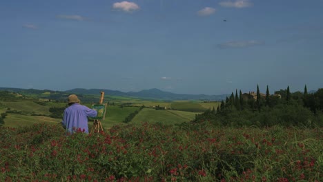 An-elderly-female-painter-is-painting-the-natural-beauty-of-the-famous-Val-d'Orcia,-also-known-as-the-Orcia-valley-as-a-painting,-standing-in-a-field-of-flowers