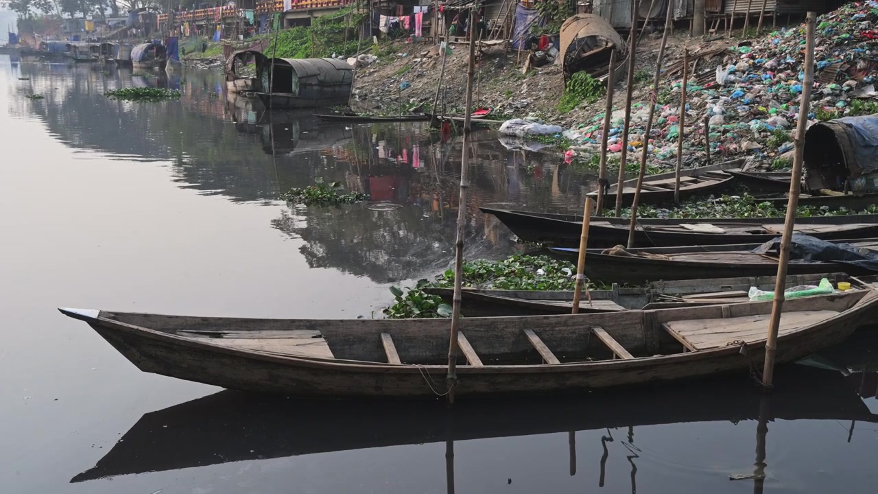 Barche di legno si riflettono su acqua calma e inquinata accanto a un'enorme pila di rifiuti di plastica in una baraccopoli di Dhaka, Bangladesh, documentando un danno ambientale estremo e lo stile di vita povero della comunità fluviale.