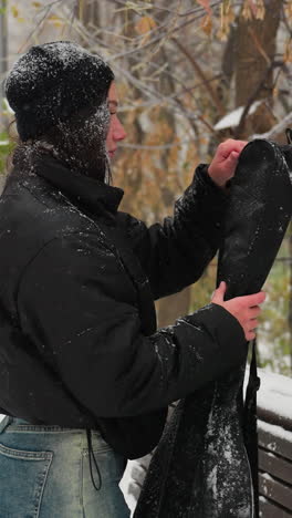 vista lateral de una mujer quitando el paquete de guitarra, colocándolo en un banco cubierto de nieve, y abriéndolo mientras la nieve cae suavemente en un tranquilo parque de invierno, rodeado de árboles helados