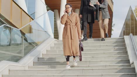 young businesswoman in elegant beige coat talking on mobile phone while going down the stairs, behind two coworkers talk