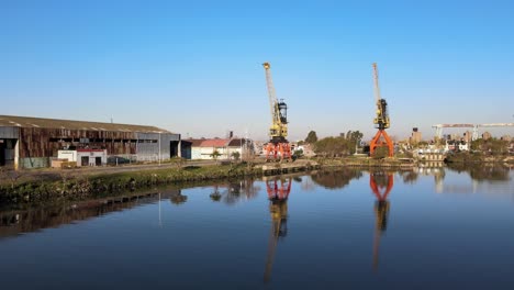 4K-Aerial-Drone-Footage-of-Two-Large-Cranes-Along-the-Water-Edge-with-Clear-Reflections-Next-to-a-Warehouse-in-Buenos-Aires-La-Boca,-Argentina