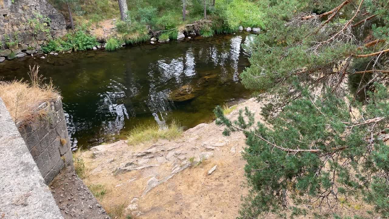 Filmen in slow motion en met een verhoogde camera de loop van de rivier de Tormes in Ávila, terwijl deze door de hoge bergen en over een stenen brug stroomt, waar een steunbeer en weelderige vegetatie verschijnen.