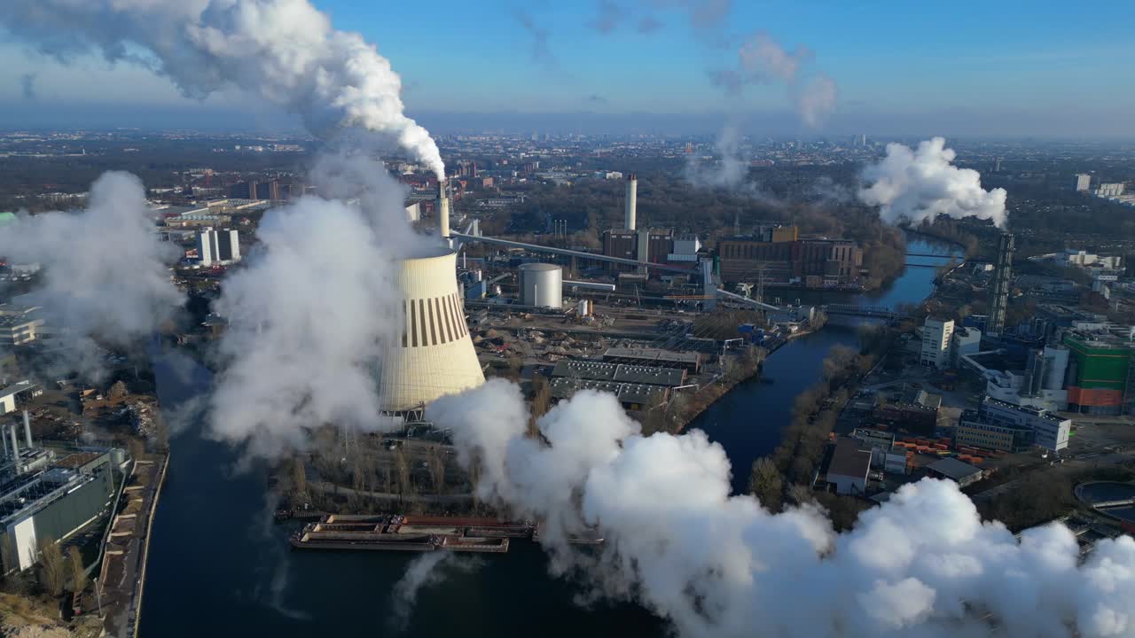 Aerial view of a thermal power plant emitting smoke on a sunny day, with a river and a city in the background. Unbelievable aerial view flight fly reverse drone