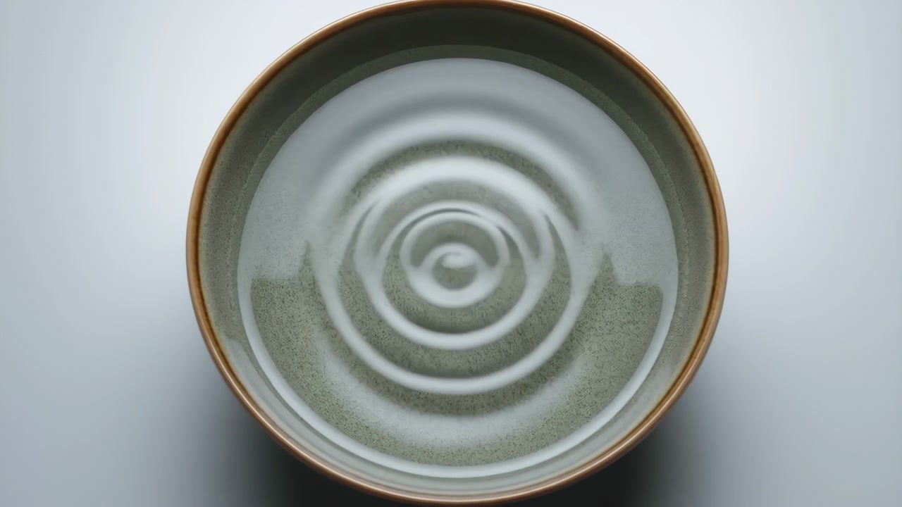 平らな表面に水滴が衝突し、穏やかで静かな真上からの視点で波紋が外側に広がる様子。グレーと白のトーンで、自然と科学を表現したシーン。