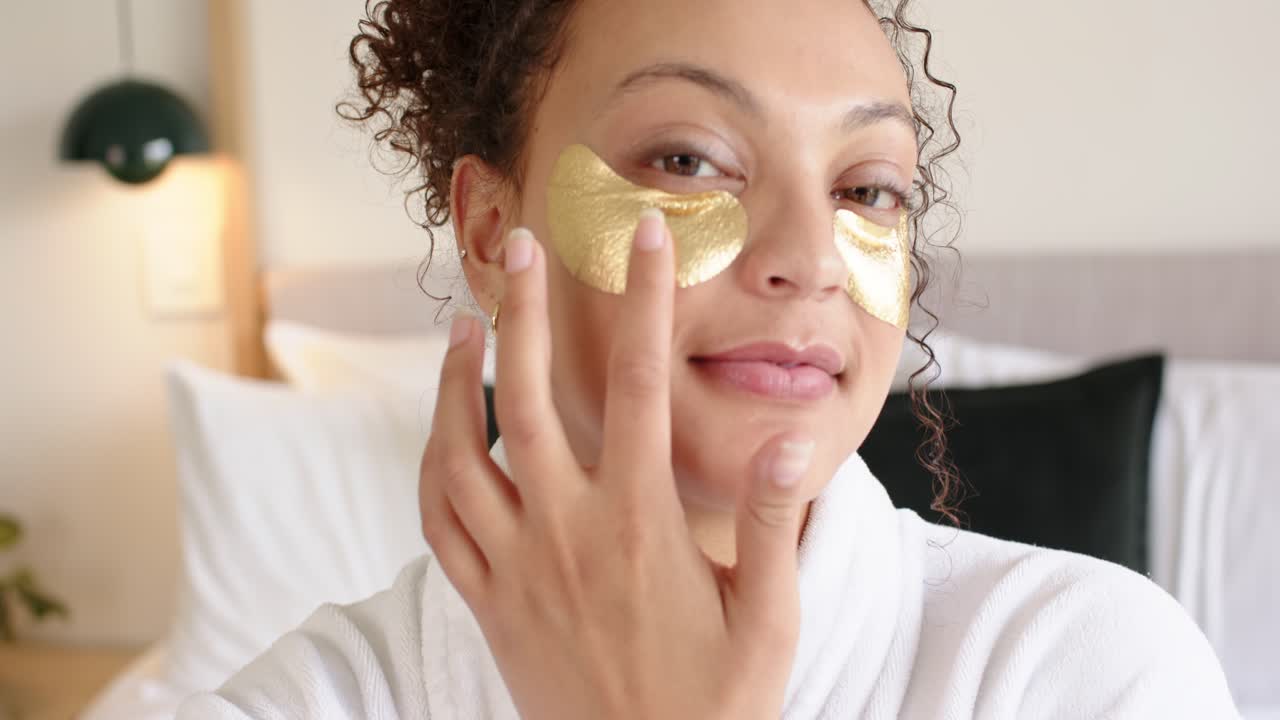 Mulher afro-americana adulta abrindo os olhos, alisando adesivos dourados para os olhos, de roupão na cama para cuidados com a pele.